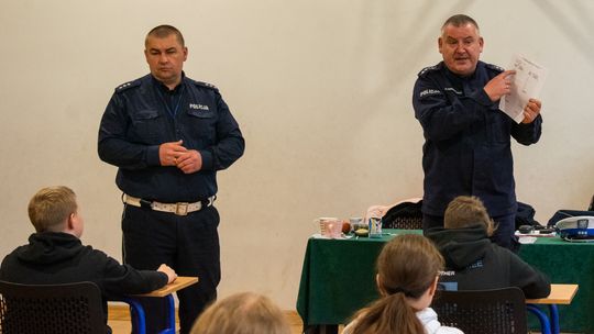 dwaj policjanci w mundurach stoją przed dziećmi siedzącymi w szkolnych ławkach